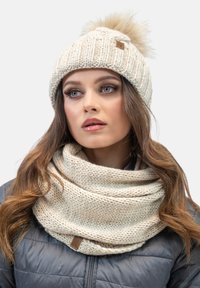 Gorro de punto de color crema con un pompón de pelo y una etiqueta de cuero marrón, acompañado de una bufanda de color crema a juego con un patrón texturizado.