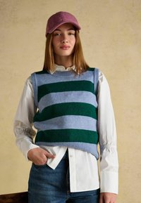 Megzta liemenė, turinti žalias ir šviesiai mėlynas juosteles, vilkima virš baltos marškinėlių su apykakle, derinama su džinsinėmis kelnėmis ir rožine kepure.