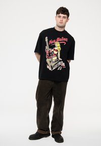 Black short-sleeve t-shirt featuring a graphic with a woman and guitar, red and cream details, paired with brown wide-leg pants and black shoes.