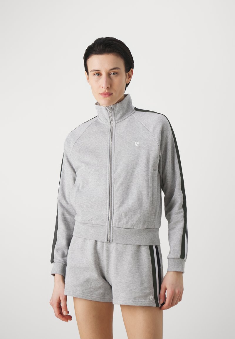 Grå hettegenser med høy krage, kontrasterende svarte sidestropper og matchende grå shorts. Myk tekstur, sporty passform, minimal merkevare.