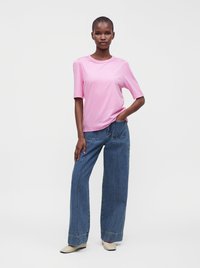 Femme portant une chemise rose clair à manches courtes, un jean bleu à jambes larges avec de grandes poches avant, et des chaussures pointues beiges, debout devant un fond uni.