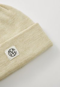 Beige strikket lue med brett og et firkantet merke som har en svart logo. Myk tekstur, klassisk design og nøytral farge.
