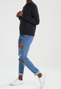 Dunkelgrauer Strickpullover mit Rundhalsausschnitt, kombiniert mit hellblauen, verwaschenen Jeans und weißen Sneakers, die große beschädigte Stellen an den Beinen aufweisen.