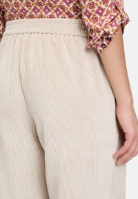 Beige corduroy-housut, joissa on joustava vyötärö, ja joissa on pystysuora ribbitekstuuri. Lähikuva, jossa käsi on lepäämässä kankaan päällä.