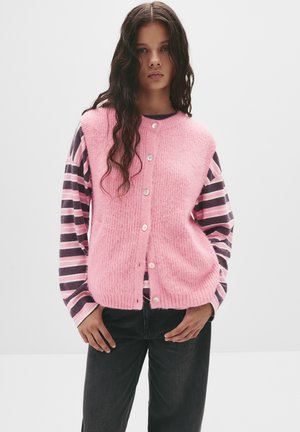 Gilet lavorato a maglia rosa con superficie testurizzata e cinque bottoni, indossato sopra una camicia a maniche lunghe a righe nei toni del viola e del rosa.
