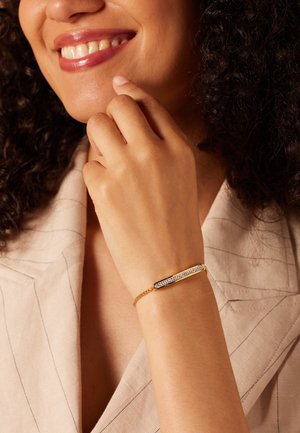 Gouden armband met een rechthoekig middenstuk met heldere stenen, gezet op een delicate ketting, gedragen om een pols met een lichte huidskleur.