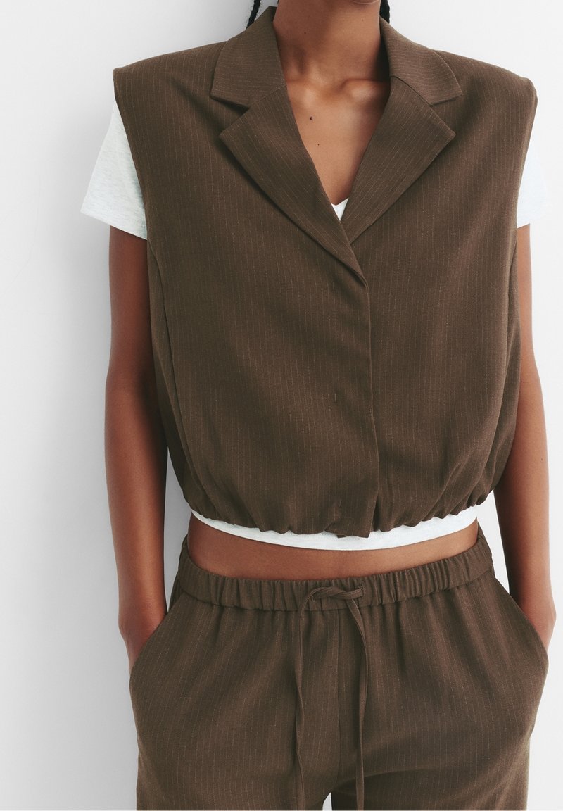 Personne portant un gilet court sans manches à rayures marron sur un t-shirt blanc et un pantalon marron à rayures assorti avec cordon, les mains dans les poches.
