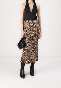 Kvinde iført en sort halterneck-top, leopardmønstret midi-nederdel, sorte støvler og holder en lille sort håndtaske foran en ensfarvet hvid baggrund.