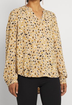 Femme portant un chemisier beige boutonné à manches longues avec un motif abstrait dispersé noir, blanc et jaune moutarde, associé à un pantalon noir.