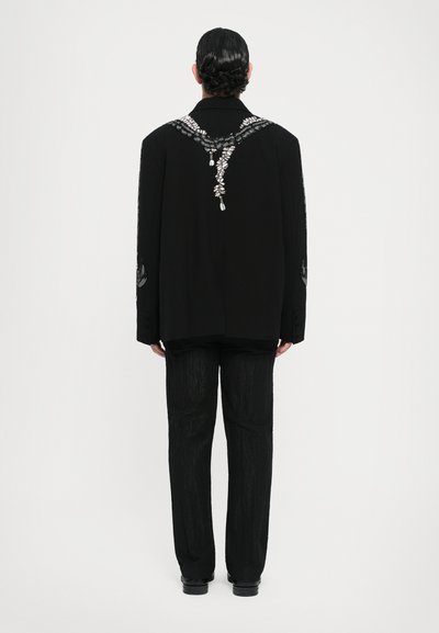 Schwarzer Blazer mit Perlenverzierungen an den Schultern und Ärmeln, kombiniert mit passenden strukturierten schwarzen Hosen. Geschmeidiger Stoff und maßgeschneiderte Passform.