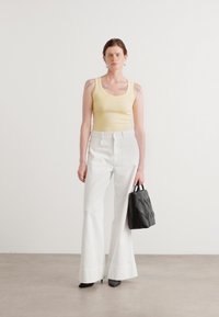 Yellow ribbed tank top, high-waisted white flared jeans, and a black textured handbag. Model wears heeled shoes, standing on a neutral background.