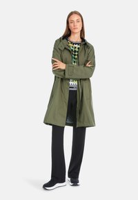 Olivgrüne wasserdichte Jacke mit Kapuze, Frontreißverschluss und schwarzen Akzenten. Getragen über einem mehrfarbigen gemusterten Oberteil und schwarzen gerippten Hosen.