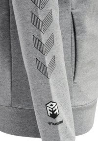 Grå sweatshirt med svarta chevronränder längs ärmen och en svart logotyp på manschetten. Tillverkad av mjukt, texturerat tyg.