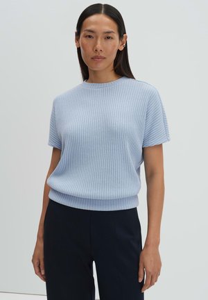 Vrouw met lang donker haar, gekleed in een lichtblauw, geribd, kort mouwtjes gebreid topje en zwarte broek, tegen een effen lichte achtergrond.