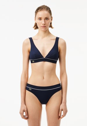 Frau trägt einen dunkelblauen Bikini mit weißem Rand und kleinen grünen Krokodil-Logos, steht vor einem schlichten weißen Hintergrund.