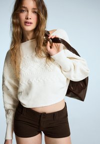 Krem pulover s pletenicama i spuštenim ramenima, uparen s tamnosmešim krojenim šortsevima. Model drži veliku smeđu kožnu torbu.