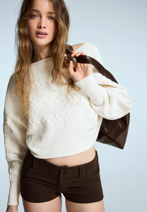 Tejelt, beszegett vállú, krém színű kötött pulóver sötétbarna, szabott rövidnadrággal párosítva. A modell egy nagyméretű barna bőr táskát tart.