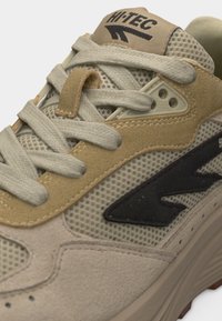 Približni plan bež i smeđih Hi-Tec tenisica s mrežastom tkaninom, preklopcima od brušene kože i svijetlosivim vezicama koje preklapaju jezik s logotipom.