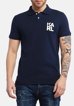 Marineblå polo skjorte lavet af bomuld med korte ærmer, en kravehals og et hvidt logo "KARL" på venstre bryst. Båret med slidte rådenim.