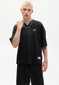 Sort mesh oversized trøje med korte ærmer, der har v-hals og hvidt logo på brystet. Bæres med matchende sorte shorts.