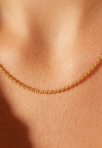 Collier en or avec des maillons entrelacés, texture lisse et éclat subtil, reposant sur une peau bronzée. Design simple et élégant.
