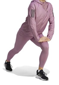 Helle lila Sportjacke mit Kapuze, versehen mit schwarzen drei Streifen- Akzenten. Kombiniert mit helllila Leggings und schwarzen Sportschuhen.