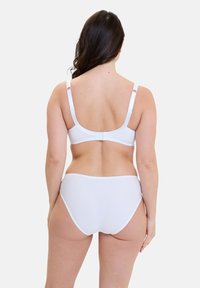 Frau, die von hinten zu sehen ist, trägt einen weißen BH und hoch taillierte Unterwäsche vor einem schlichten hellen Hintergrund.