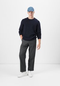 Granatowy sweter z przędzy w plamki, szare spodnie i białe sneakersy. Model nosi jasnoniebieską czapkę, stojąc na tle jednolitej białej ściany.