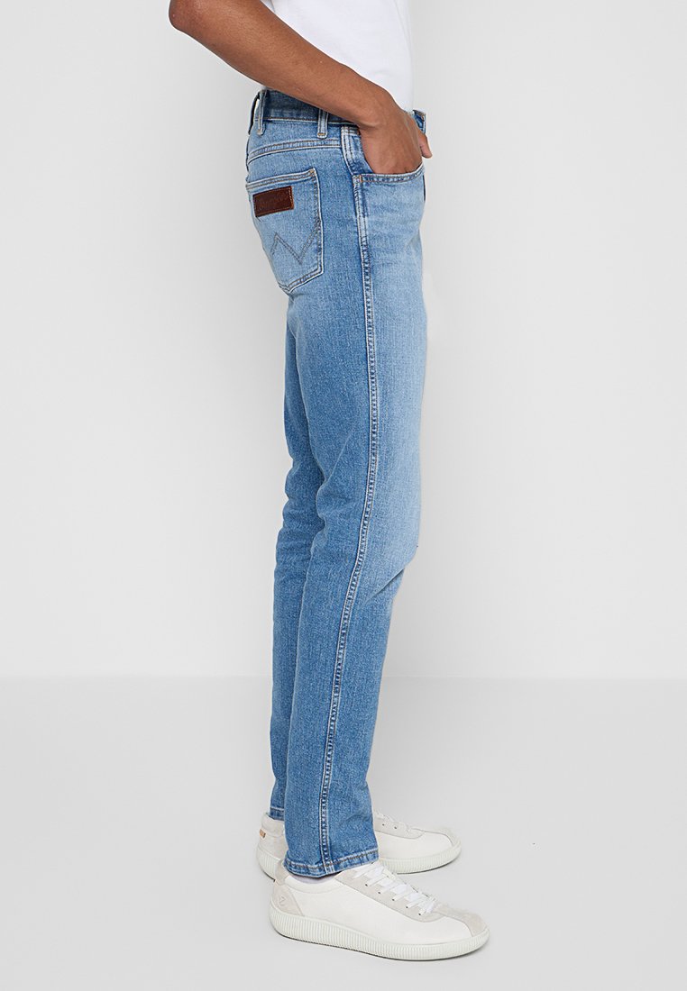 Jasnoniebieskie dżinsy o slim fit, pięć kieszeni, skórzana naszywka z tyłu oraz kontrastowe szwy. Noszone z białymi trampkami.