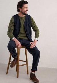 Man som sitter på en trästol, klädd i en mörkblå väst över en olivgrön tröja, mörka byxor och bruna ankelstövlar, med uppvikta ärmar och flerfärgade strumpor.