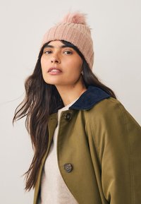 Világosrózsaszín bordázott kötött sapka bolyhos pompommal, olívazöld dzsekivel viselve, amely tengerészkék kordbársony gallérral és gombos záródással rendelkezik.