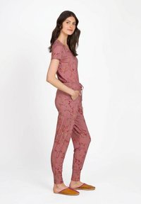 Woman standing sideways wearing a mauve floral print jumpsuit with pockets and brown slip-on shoes.