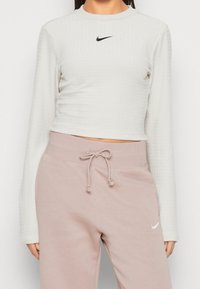 Avkortad långärmad toppen i strukturerad vit tyg med en liten svart Nike-logotyp. Kombineras med lösa rosa sweatshirtbyxor med elastisk midja.