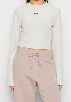 Kratztopper mit langen Ärmeln aus strukturiertem weißen Stoff und kleinem schwarzen Nike-Logo. Kombiniert mit lockeren rosa Sweatpants mit elastischem Bund.