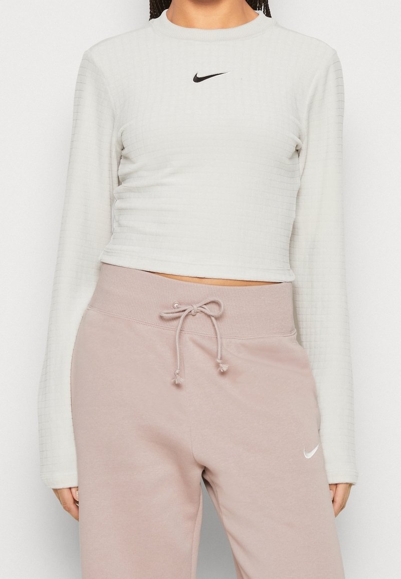 Avkortad långärmad toppen i strukturerad vit tyg med en liten svart Nike-logotyp. Kombineras med lösa rosa sweatshirtbyxor med elastisk midja.