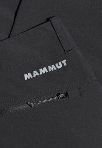 Svarta Mammut-byxor har en zip-ficka och ett strukturerat tyg. Logotypen är tydligt synlig i reflekterande bokstäver.
