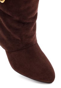 Brown suede ankle boot with a pointed toe and slouchy design. Features a gold-colored accent near the top. Smooth texture throughout.