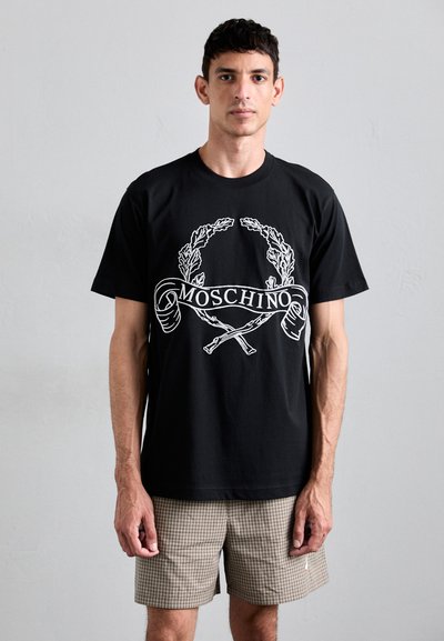 Czarna bawełniana koszulka z białym nadrukiem graficznym przedstawiającym liście laurowe oraz napisem "MOSCHINO". Model nosi szorty w kratę.