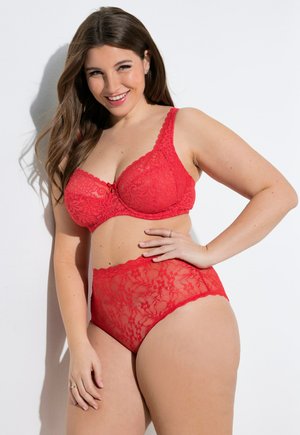 Usměvavá žena s dlouhými hnědými vlasy, oblečená v červené krajkové podprsence a kalhotkách s vysokým pasem, pózující na bílém pozadí.