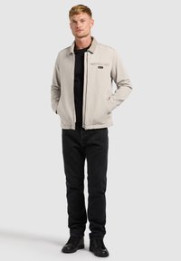 Homme portant une veste beige zippée sur un t-shirt noir, un jean noir et des baskets noires, debout les mains dans les poches sur un fond neutre.