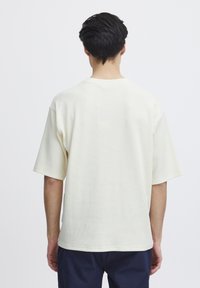 Krátký rukáv, volný crewneck tričko v barvě světlé smetany; texturovaná tkanina; má jednoduchý design bez viditelných grafik.