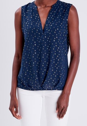 Ärmellose marineblaue Bluse mit kleinem weißem Blumenmuster, V-Ausschnitt und gedrehtem Saum, getragen mit weißen Hosen.