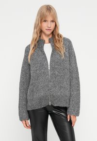 Cardigan gris tricoté avec une fermeture éclair, texture côtelée, manches larges et col haut, porté sur un haut blanc et un pantalon noir.