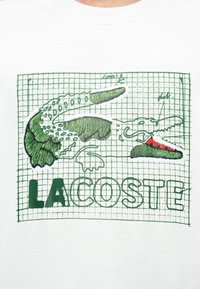 T-shirt bianca con un grafico di coccodrillo ricamato in verde con motivo a griglia e testo "LACOSTE" in lettere verdi in grassetto nella parte inferiore.