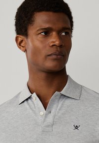 Muž s krátkymi kučeravými vlasmi, oblečený v svetlosivej polokošeli s dvoma gombíkmi a malým logom skrížených vesiel na hrudi.