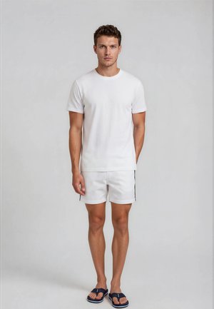 Man standing against plain background wearing white t-shirt, white shorts with black stripe, and navy flip-flops, facing forward with arms relaxed.