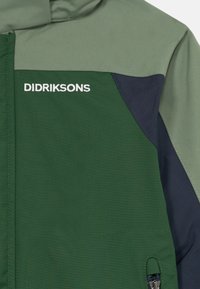 Roheline, heleoliiv ja tumesinine veekindel jope, millel on valge "DIDRIKSONS" logo ja esiküljel lukuga tasku.