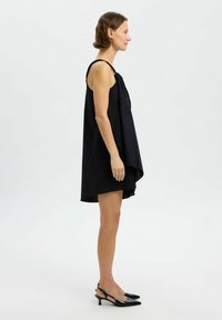 Mujer con un vestido negro asimétrico sin mangas y zapatos slingback de tacón bajo negros, de perfil sobre un fondo blanco.
