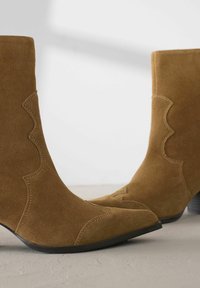 Ruske suede gležnjače s oštrom nosom, valovitim detaljima i niskim složenim potplatom na teksturiranoj površini.