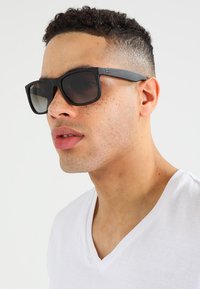 Ochelari de soare negri, rectangulari, cu lentile întunecate, cadru din plastic și branding subtil pe temple. Modelul poartă un tricou alb cu guler în V.